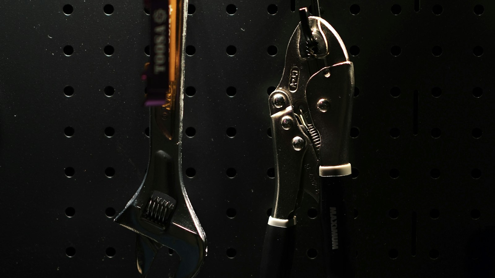 An adjustable wrench and locking pliers hanging on a dark pegboard, with empty hooks around them