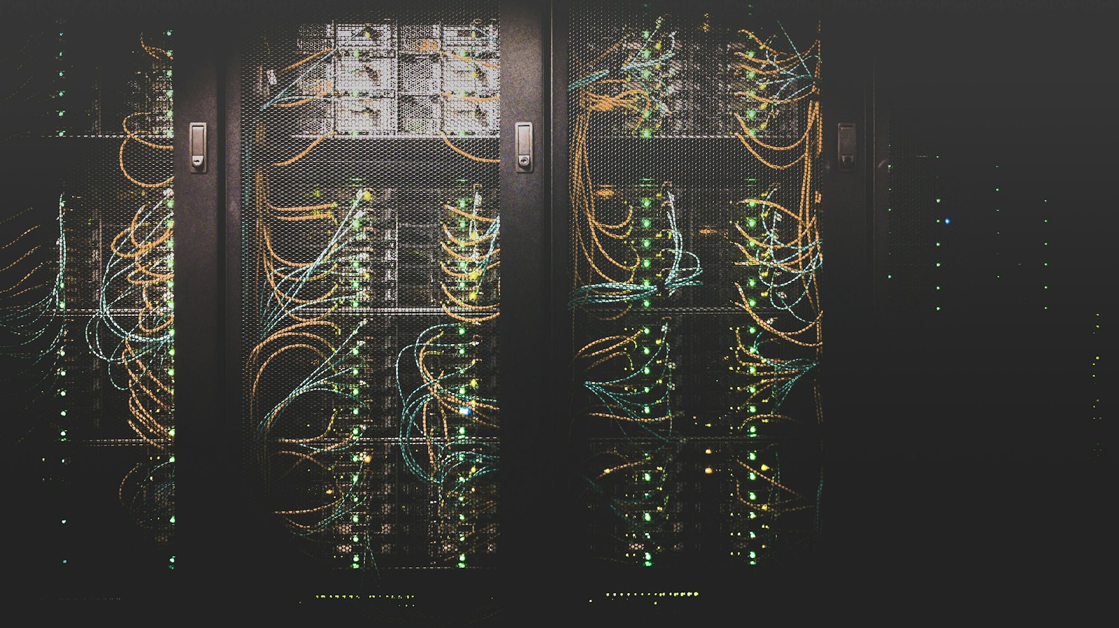 Rows of dark server racks in a data centre, threaded with orange and teal patch cables and lit by green status LEDs